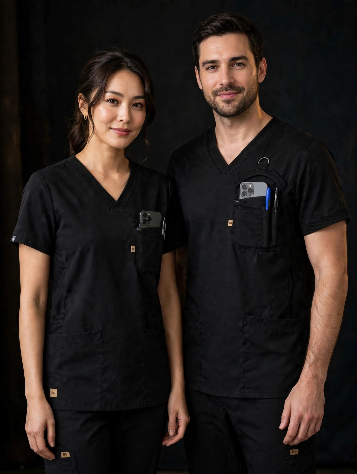 Two models wearing black BG Scrubs — the man's top carries the signature arch retention pocket with D-ring, phone and pens; both tops show the ribbed V-neck and BG tags